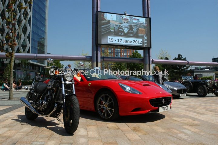 A motorcycle parked on the side of a street - Pistonheads - The image shows an urban setting with a focus on a red Ferrari sports car parked on a brick pavement next to a sidewalk. Behind the car on what appears to be a promotional platform, there are multiple other cars displayed, including some classic and modern models. Above the platform, a large billboard screen is prominently displaying the dates "12 - 16 JUNE 2012" and the word "FERRARI," suggesting an exhibition or event related to the brand. In the background, there are tall buildings, indicating that this scene is taking place in a city environment. The presence of other people and items suggests a public or communal space, possibly accessed from the platform on which the Ferrari sports car is parked.