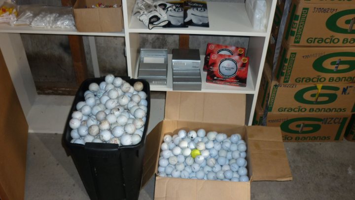 Golf - how bad as a beginner? - Page 1 - Sports - PistonHeads - The image captures an indoor setting featuring a collection of boxes and baskets organizing on two white shelves. On the left side of the image, there's a large black plastic bin overflowing with white sports balls. A yellow bird is perched atop this bin. Next to it on the floor, an open cardboard box displays a small number of these balls. Further right, another white shelf holds a full box of white items with a green label, and a red item with a white label. In the background, behind the shelves, there's a glimpse of a wall with a window, and cardboard boxes are stacked against the wall, filling the top of the image from left to right. The lighting in the room comes in from the window, casting a warm glow.