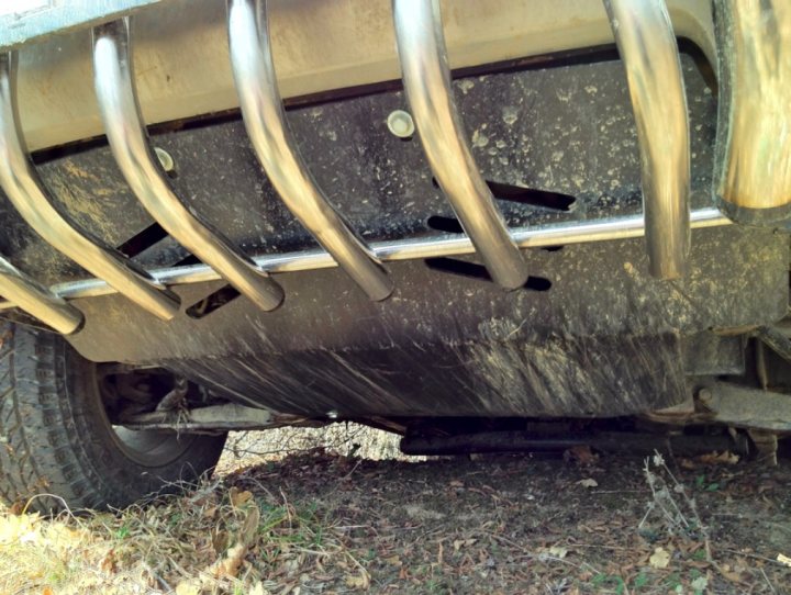 Pistonheads - The image shows a scene under a vehicle, highlighting an up-close perspective of its rear bumper and part of the chassis. The vehicle seems to be a car. The perspective is from a distance, positioned either directly under the vehicle or at a slight angle. The ground appears to be wet, and there is some vegetation in the foreground, consisting of patches of grass and fallen leaves. There is no visible text or distinctive branding in this image.