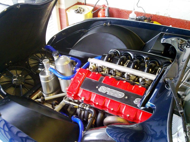 Any pics of silicone hoses for Tuscan, black or blue? - Page 1 - Speed Six Engine - PistonHeads - The image provides a view of the engine compartment of a motorcycle. The engine is painted in a blend of red and black colors. Several tubes and hoses are visible, connecting different parts of the engine. There is a prominent metal cooler placed on the side of the engine. The engine is mounted in a black frame, providing structural support to the engine compartment.