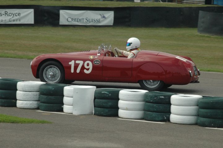 A red and white fire hydrant sitting on the side of a road - Pistonheads - The image depicts an exciting scene from a slalom auto race. At the center of the image, a vintage red race car, with the number 179 prominently displayed on its side, is skillfully navigating a track. The driver, donned in a helmet and a matching white racing suit, is in the midst of dynamically leaning into a turn around a barrier. The barrier itself is lined with seven stacks of tires, indicating the challenging nature of the race track. In the background, a black sign with the words "Credit Suisse" is visible, possibly indicating the event's sponsors or the location of the event.