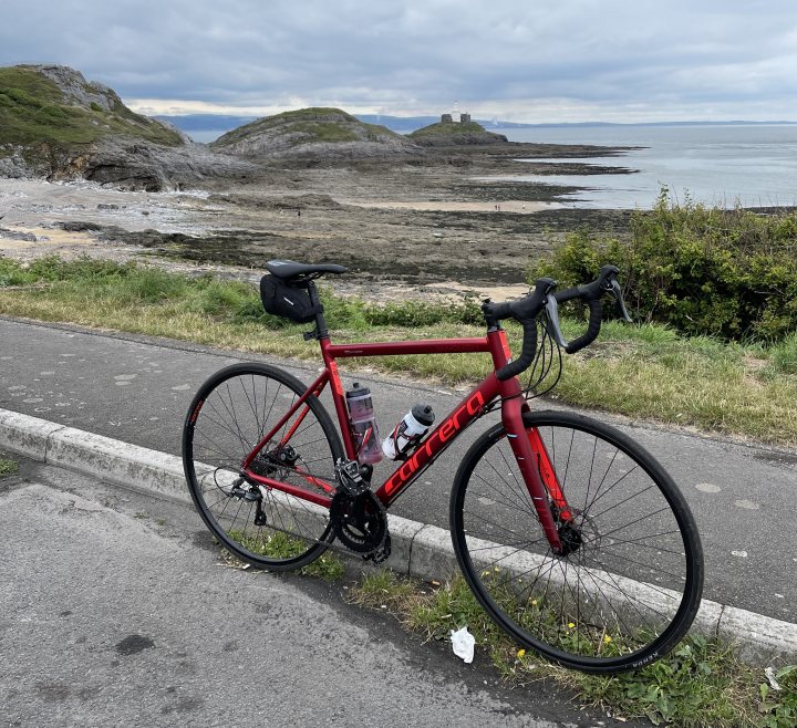 The "Photos From Today's Ride" thread. (Vol. 2) - Page 26 - Pedal Powered - PistonHeads UK - The image features a road bicycle parked on the side of a road. It's a clear day with some clouds in the sky, suggesting it might be early morning or late afternoon. The bicycle is leaning against a curb and appears to be of a high-quality build, possibly used for long-distance rides given its robust frame and multiple water bottle mounts. In the background, there's a scenic view with a coastline, where the road seems to follow along the shore, with small hills or dunes nearby. The presence of this bicycle suggests that someone has taken a break from their ride to enjoy the picturesque view.