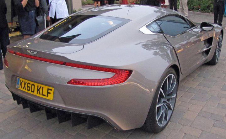 Pics from Aston event, Monterey - Page 1 - Aston Martin - PistonHeads - The image showcases a sleek gray sports car parked on a brick sidewalk near the street. The car's front is facing directly towards the camera, revealing the elegant design of its hood and the rear. The rear section features a unique lighting setup with multiple red lights in a horizontal line which adds to the car's distinctive look. The bricks under the car contrast with its shiny metallic finish, while people and other elements of the scene can be seen in the background, suggesting that this scene might be part of a car show or a busy city street.