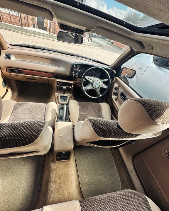 Pistonheads - The image shows the interior of a car, focusing on the front passenger seat and dashboard. This car appears to be a luxury vehicle with tan leather upholstery and wood grain inserts. The steering wheel is visible, indicating that the car is right-handed drive. The dashboard includes a radio or stereo system. The window is partially open, allowing light into the cabin. There's a sunroof in the ceiling that's currently closed.