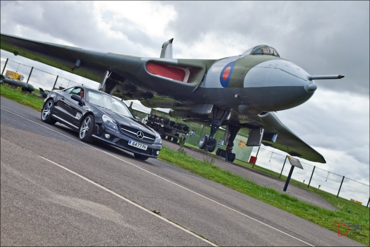 So what have you done with your Aston today? - Page 226 - Aston Martin - PistonHeads - This image captures a striking scene where an olive green fighter jet with a distinctive Union Jack on its tail is parked on a runway. In front of the jet, a black Mercedes sports car is parked, seemingly ready for a high-speed dash. Surrounding the jet and car are a few street lights and fences, suggesting that this is a military airfield or aviation museum. The sky above is filled with dark, ominous clouds, creating a dramatic and provocative atmosphere. The juxtaposition of the fast-moving jet and the luxurious car on the runway creates a sharp contrast between the contrast and the calm, quiet environment of the airfield.