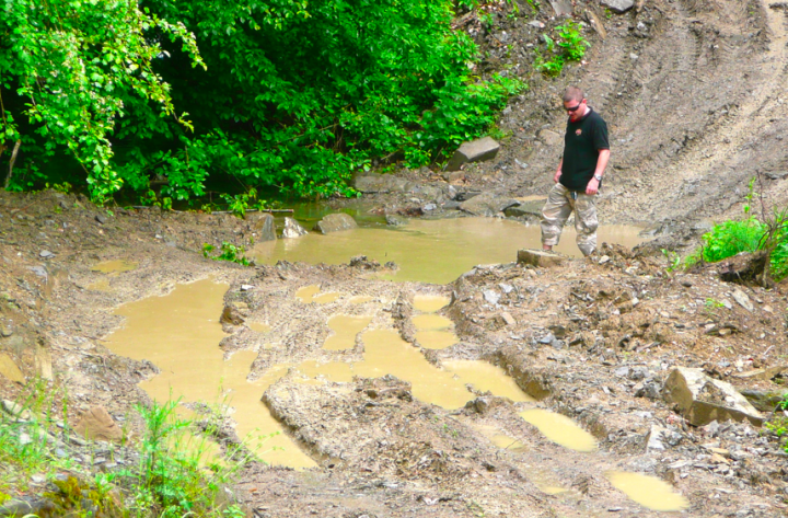 Little overnight trip to the Balkan range - Page 1 - Off Road - PistonHeads - The image shows a man standing amidst mud and water, near a path that appears to be a trampled dirt trail. He is in the middle of a muddy parking lot with waterlogged areas. The man himself is dressed in attire that includes sunglasses, a black shirt, and shorts, suggesting a possible outdoor activity or work. His posture and the fact that he is half-submerged in the mud indicate he might be involved in the cleanup or construction work, given the sparse vegetation around him. The surrounding area seems to be in a natural or semi-urban setting, with trees and rocks visible in the background, adding a rustic charm to the scene.