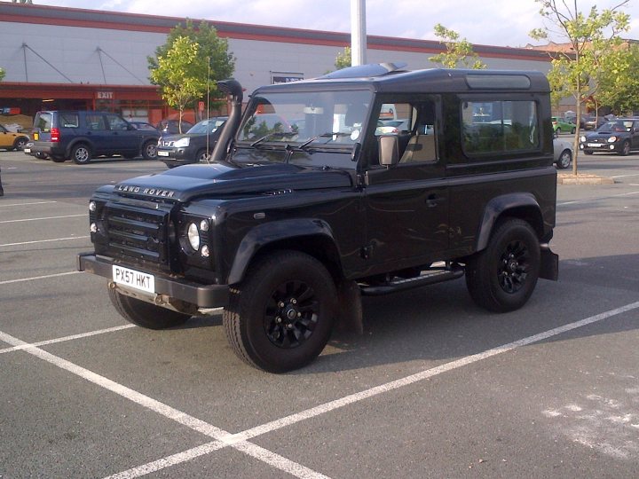 A white truck parked in a parking lot - Pistonheads - The image shows a black, four-door Range Rover parked in a public parking area. The vehicle is positioned at an angle to the viewer, allowing a clear view of its front and side. The parking lot is lined with white boundary markings. In the background, there are other cars parked and a building with a red exterior. The sky appears to be overcast, contributing to the muted lighting in the scene. The overall appearance of the vehicle and the setting suggest this photo might have been taken in an urban or commercial setting.