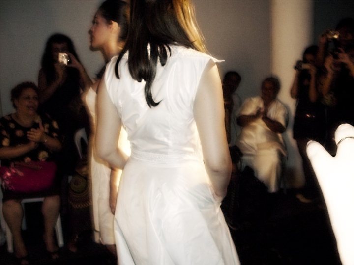 Ncego - The image captures a moment of performance, where one woman is the central focus, captured in mid-stride against a blurred background. She is wearing a white dress and walking away from the camera with her back to the viewer, allowing a sense of motion in the frame. In the background, other people are seated and standing, observing the performance, with elements of a ceremony suggested by the presence of a chair and a handbag. The lighting highlights the central figure, creating a dynamic contrast against a softly lit audience.