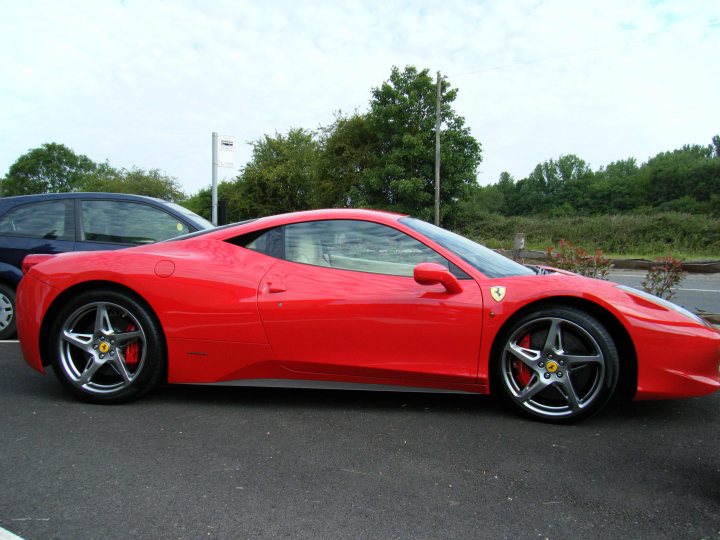 Breakfast Meeting, The Hare, Roxwell 4th June - Page 1 - Kent & Essex - PistonHeads - The image features a red Ferrari parked on the side of a road. The car is positioned at a slight angle that allows a clear view of its profile and front wheel. The setting appears to be during the day with natural light illuminating the vehicle, highlighting its sleek design. In the background, there are trees, a building, and another car, suggesting a suburban or urban environment. The road seems quiet and open, with no visible traffic or pedestrians. The Ferrari's color contrasts sharply with the more muted tones of the surrounding area, drawing attention to it as the main subject of the image.