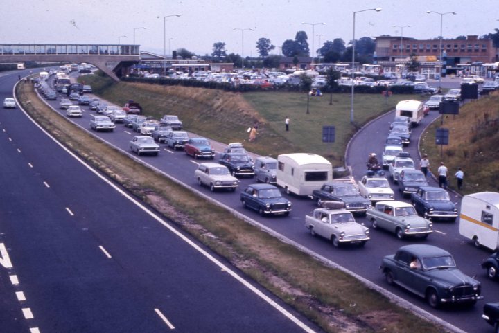 A 'period' classics pictures thread (Mk II) - Page 44 - Classic Cars and Yesterday's Heroes - PistonHeads - The image presents a bustling highway scene, viewed from an elevated perspective. Numerous cars of varying models are captured in motion, driving along the road. A few trucks can also be seen amidst the vehicular traffic. The highway appears to be well-maintained with clear lane markings. There's no sign of pedestrians on the sidewalks that run parallel to the highway, suggesting this might be a less populated area or time of day when foot traffic is minimal. The overall scene paints a picture of everyday life and movement in a modern urban setting.