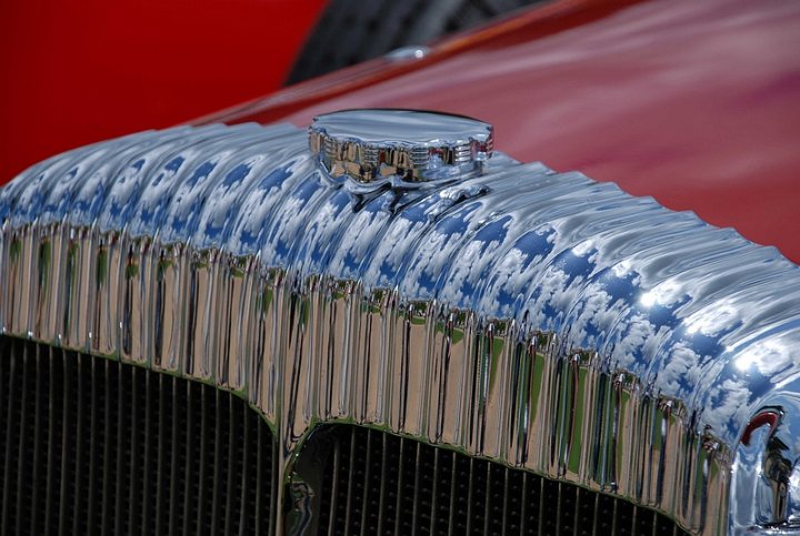 GRRC Lunch Club - Page 1 - Goodwood Events - PistonHeads - The image captures a close-up view of the hood of a vintage car. The car features a vibrant red body with a shiny chrome bumper and grill. The grill is intricately designed, with large vertical bars that reflect the clear blue sky. The front of the car is detailed and polished, suggesting it is well-maintained and likely a collector's item. The focus of the image is the shiny chrome, which contrasts with the matte red paint and the blue sky in the background.