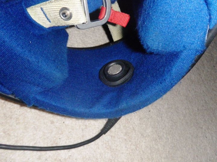 Pistonheads - The image shows a close-up of the underside of a blue car seat. A strap attachment is visible, featuring a clasp with a baby monkey shaped red tag. The car seat is resting on a carpeted floor and a section of the webbing from a child's harness is also partially visible in the foreground. There's a hint of another object to the left side of the image.