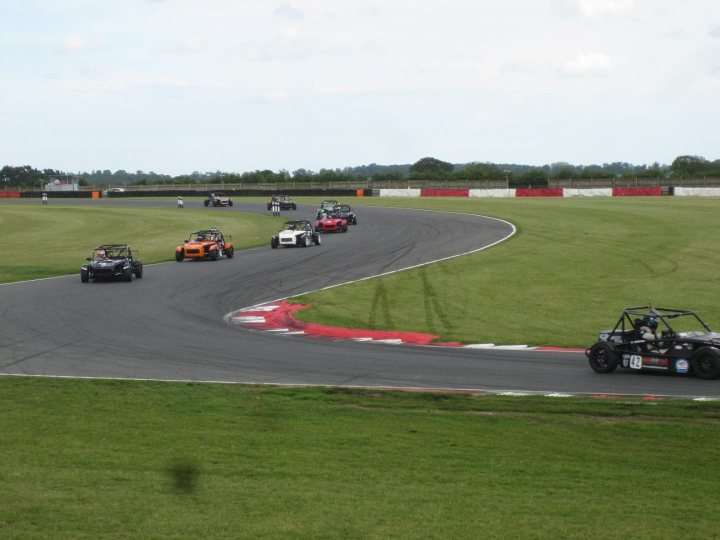 750MC Roadsports - Bank Holiday Monday - Snetterton - Page 1 - UK Club Motorsport - PistonHeads - This is an aerial shot of a race circuit with several cars on a curved track. The vehicles are driving through the circuit, some of them leading ahead, capturing the action of the race in progress. The track is paved and it appears that the cars are racing vehicles designed for speed and maneuverability. The surrounding area is green, suggesting it might be spring or summer, and there are flags lining the track, which adds to the competitive atmosphere of the race.