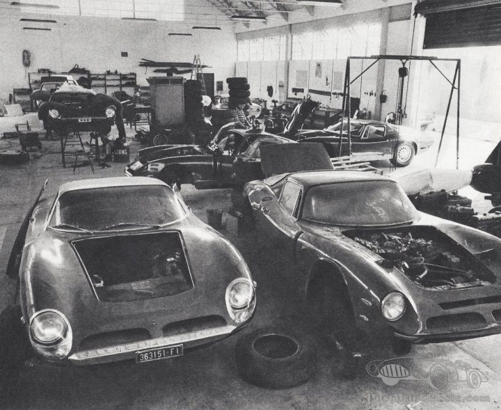 A 'period' classics pictures thread (Mk III) - Page 41 - Classic Cars and Yesterday's Heroes - PistonHeads UK - The image is a black and white photograph depicting an interior space that appears to be a vehicle workshop or garage. In the foreground, two vintage sports cars are parked side by side. One car has its hood open, revealing its engine, while the other car's hood remains closed. The cars appear well-maintained and polished, indicative of a setting where they may be restored or preserved.

Surrounding the cars, there is an assortment of vehicles and parts, including another vintage sports car in the background and various mechanical components scattered around the floor. On the right side of the image, there's a person standing among the vehicles, possibly working on them. The overall atmosphere suggests a workshop or garage dedicated to classic or vintage cars.

The setting is characterized by high ceilings and ample natural light coming from windows not visible in this portion of the image. The walls are bare, allowing the focus to remain on the vehicles themselves and their immediate surroundings. There is no text present in the image, and the style of the photograph conveys a sense of history or nostalgia, possibly implying that these cars may be of great value or significance to their owners or enthusiasts.