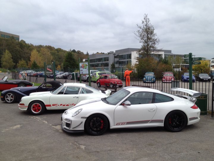 Just picked up a 4.0RS - Page 3 - 911/Carrera GT - PistonHeads - The image captures a gathering of classic and sporty cars parked in a spacious lot. In the foreground, a pristine white Porsche 911 GT3 RS parked on the center stands out with its red wheels and distinctive rear wing. To its right, a green Porsche 996 GT2RS is parked along with a blue Porsche Cayman parked a bit further back. The vehicles have a sleek design and appear to be well-maintained. In the background, the lot is lined with parking spaces, some of which are occupied by random vehicles. The setting suggests this might be a car meet or a special day for classic car enthusiasts.