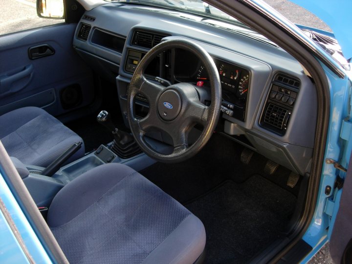 Pistonheads Ford Sierra - The image depicts the interior of a gray Ford car. A black steering wheel is positioned in the center of the image, and a matching black seatbelt is visible to the left of the front passenger seat. A blue seatbelt is situated to the right of the steering wheel. The car has a two-door design, and the open driver's side door provides a clear view of the vehicle's interior. The steering wheel is equipped with various buttons and controls, typical of Ford vehicles. The paint condition of the car appears to be in good condition, with the gray bodywork neatly painted.