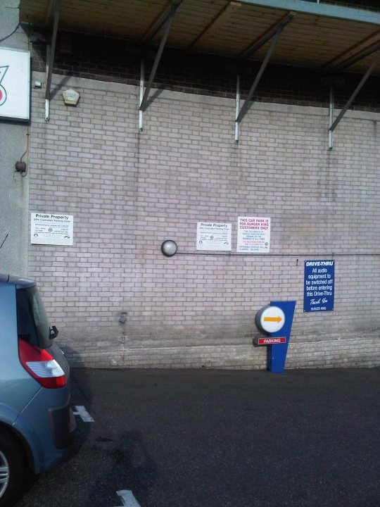 Pistonheads - The image shows a parking lot scene against a brick wall. There is a silver car parked close to the wall on the left side. On the wall, there are three white signs with text and a red and white arrow sign pointing to the right. There is also another sign above the brick wall. The sun is shining on the wall, highlighting the signs and brick texture.