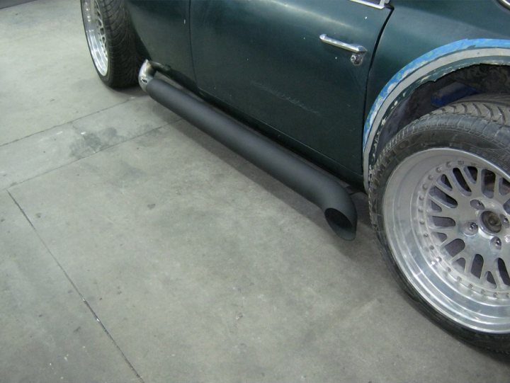 3428TM Build Log - Page 11 - Classics - PistonHeads - The image shows the side of a car equipped with a large exhaust pipe. The car is painted green and features a silver rim on the wheel on the side. The flooring appears to be concrete, and other vehicles can be seen in the background. The photograph appears to be taken indoors or in a contained area, given the uniform surface and the presence of a line or seam running horizontally across the photo.