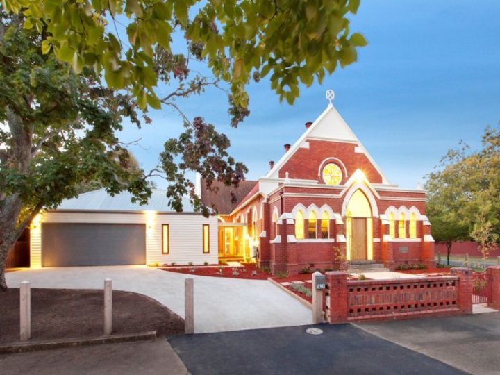 melbourne & other frequently asked questions - Page 1 - Australia - PistonHeads - The image shows a two-story building with a distinctive architectural style, characterized by a large central arch, a prominent round window on the upper level, and a peaked roof. The building appears to be a mix of ornate and classical elements, potentially a church or a similarly meaningful community structure. In front of the building, there is a well-maintained yard, and the structure is illuminated by outdoor lighting, suggesting it is either early morning or late evening. A sidewalk leads up to the entrance, also lit up, and there's a glimpse of a red brick wall in the background. The sky is clear and blue, indicating fair weather.