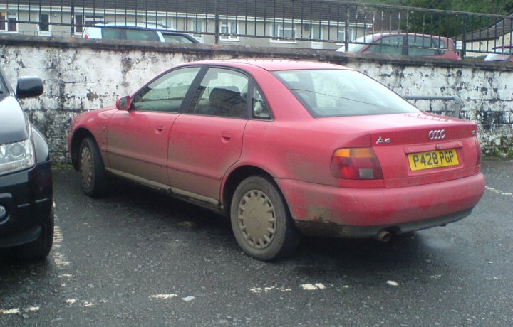 The BAD PARKING thread [vol3] - Page 267 - General Gassing - PistonHeads - The image features a red Audi, a smaller car model, parked in an outdoor parking lot or lottery office. The car has a distinct European license plate and is positioned next to a white brick wall. The ground appears wet, suggesting rainfall may have recently occurred. The car's passenger side window is closed, and the vehicle's overall condition appears to be moderately worn, with noticeable mud splatters on the rear quarter panel vehicle.