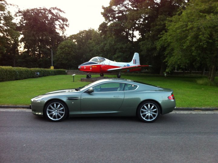 Show us your DB9  - Page 3 - Aston Martin - PistonHeads - In the image, a sleek silver Aston Martin is parked on a road, adjacent to a vibrant red and white airplane. The airplane, parked on concrete, has a distinct vintage design. The road is surrounded by lush greenery, and the plane and car are parked in a gravel area by the side of the road. The overall scene suggests a setting of luxury and exclusivity, possibly hinting at a private airfield or airport.