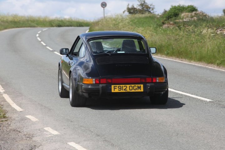 Caution! 911 and CGT content. No residual value chat! - Page 2 - 911/Carrera GT - PistonHeads - In the image, a classic black BMW is captured in mid-drive on a country road. The car is angled slightly to the right, suggesting its direction of movement. The surroundings are lush, with verdant grass on either side of the road, hinting at a rural or suburban location. The condition of the road is good, with marked center and edge lines, indicating it's likely a well-traveled route. The sky above is a clear blue with a few scattered clouds, suggesting a sunny day.