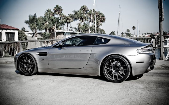 Wheel business..... - Page 1 - Aston Martin - PistonHeads - The image features a shiny silver sports car parked on a street. The car has a sleek design with a sunroof and large, aftermarket wheels. The car is facing towards the right side of the photo, and there are palm trees visible in the background, suggesting the photo might have been taken in a coastal area or a place with a warm climate. There's a chain-link fence next to the car, which adds a gritty feel to the scene. The car's sleek design contrasts with the industrial look of the gate and the fence in the background.