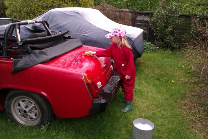 MG Midget - Just get it finished !! - Page 1 - MG - PistonHeads - The image shows a young girl washing a red car. She is wearing a white baseball cap with red polka dots, a red jumpsuit, and green rain boots. Her hands hold a yellow sponge while she splashes water on the car, which is parked on a lush green lawn. Behind the car, there's a white tarps and a wooden fence. The overall scene suggests a casual day out where the girl is engaged in practical chores.