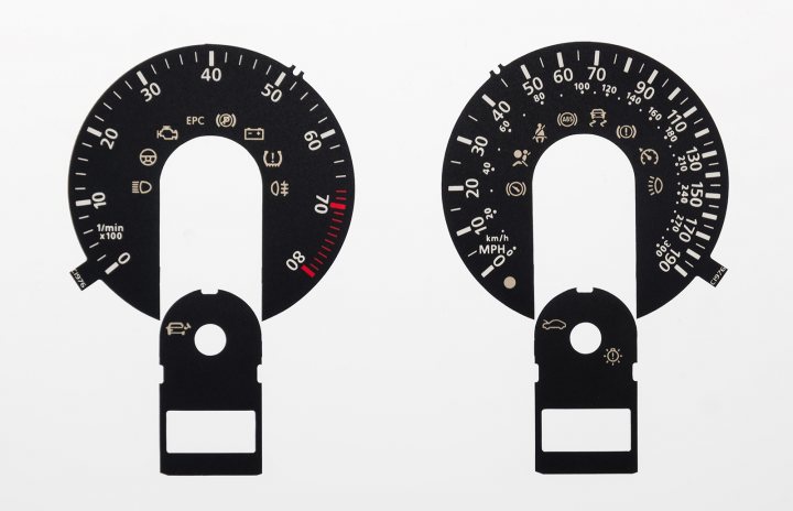 The image displays two automotive-themed items against a white background. On the left, there is a speedometer gauge face, with red and yellow needles indicating different speeds. This gauge features black numbers and markings, along with the words "SPEEDOMETER" inscribed at the top.

On the right side of the image, there is a circular device that appears to be a steering wheel cover or a speedometer-like accessory for a model train. It has a black background with white and red numbers around its circumference, representing different speeds or settings. The center of this item has a red needle and a small gold emblem in the middle.

Both items are likely meant to represent speed measurement devices, one for a vehicle and the other for a model train, as suggested by their design.