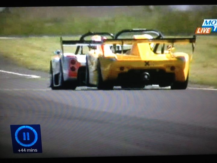 Flying - Page 1 - Radical - PistonHeads - The image captures an intense moment of a head-to-head race between two race cars. They are on a track, evident by the white stripes on the asphalt surface. The car in the foreground is predominantly white and red. In contrast, the car in the background is a bright yellow. The cameras are tracking these vehicles from a rear side angle, providing a dynamic race perspective. The image also contains playground equipment, suggesting that there might be a play area nearby, which brings an unexpected juxtaposition to the serious nature of the car race.