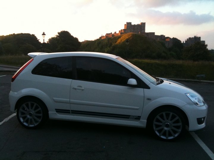 Pistonheads - The image presents a scene at sunrise with a white Volkswagen compact car parked by a castle-like structure. The car is sleek and modern, with a distinctive design featuring alloy wheels, a spoiler, and chrome accents. The castle, possibly a historical or ancient building, indicated by its grandeur and robust walls, provides a striking contrast to the small, sporty vehicle. The parking lot appears to be spacious and empty, except for the Volkswagen, which takes up most of the frame. The camera angle seems to be from the perspective of someone standing behind the car, looking out toward the distant castle.