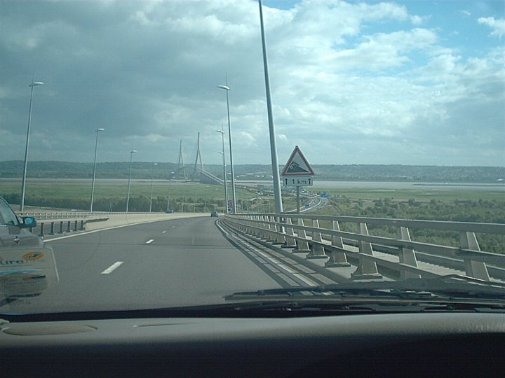 Pistonheads Rochelle Lehavre - The image captures a wide, multi-lane road at a distance, with a few vehicles visible, including a truck. In the upper right corner, there's a triangular traffic sign with circular symbols warning of potential unfavorable weather or road conditions. The sign is red with a line across it. The sky in the upper left corner appears overcast with a mix of clouds and bright sunshine, suggesting a dynamic weather situation.