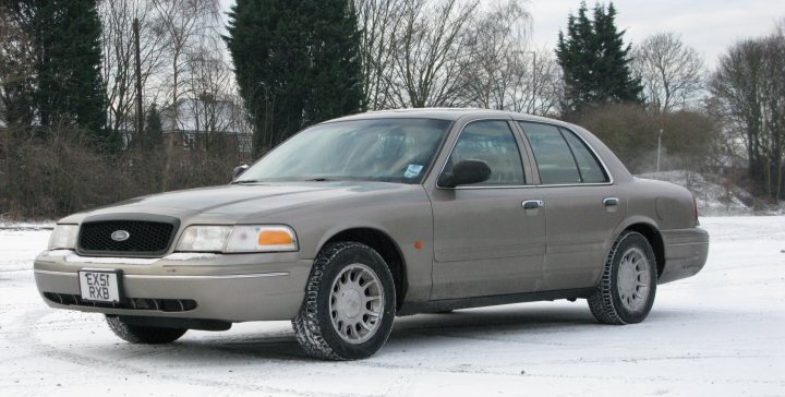 Yank Tank Pistonheads - The image shows a grey sedan parked on a snow-covered road. It appears to be a Ford, given the shape and design. The car has tinted windows and is equipped with standard steel rims. The image itself is a bit grainy, suggesting it might have been taken in low light or during a period of intense cold, which is further indicated by the snow-covered surroundings, including trees and what appears to be a distant building hidden by the snowfall. The car is positioned at an angle, with a clear view of the front grille and headlights, allowing the license plate to be visible.