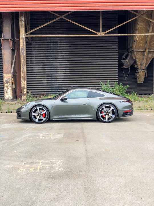 Aventurine Green - Page 2 - Boxster/Cayman - PistonHeads - The image shows a gray sports car parked in front of a large, industrial building. The car features a sleek design with prominent wheels and a sporty rear wing. The vehicle is positioned on the side of what appears to be a concrete surface or parking lot. The building has rusted metal panels and a large door frame visible in the background. There are no people present in the image, focusing attention on the car and the industrial setting.