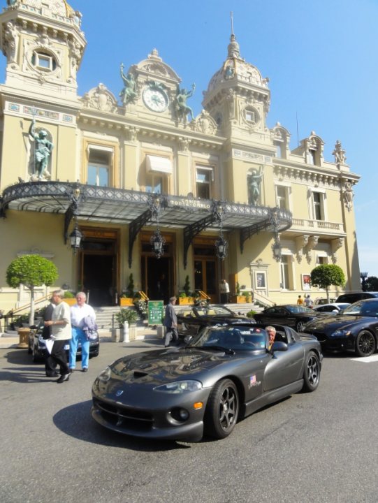 Celebrating 20 years of ... - Page 1 - Vipers - PistonHeads - In the image, a group of people are gathered on a sidewalk in front of an elegant European-style building. The building, tall and grand, has a clock tower and is decorated with statues and intricate details. Below the building, several cars are waiting at a stoplight, including a black sports car that is closest to us, with two occupants inside. The scene is cordoned off by security guards, and the overall atmosphere suggests a formal or ceremonial event.