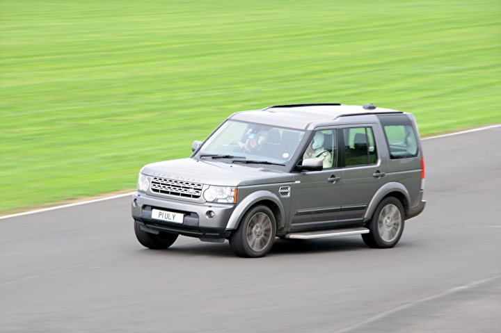 Unusual track day cars? - Page 1 - Track Days - PistonHeads - The image shows a gray SUV driving on a track. The vehicle is characterized by a rugged and sturdy build, with a large body and raised ground clearance. Its front grille is prominent and designed with a series of horizontal bars. The vehicle is equipped with tinted windows and five-spoke silver alloy wheels. Two individuals are visible inside the car, possibly the driver and a passenger, and they are focused on the road ahead. The setting appears to be a race track or a special off-road event, given the road conditions and the vehicle's appearance.