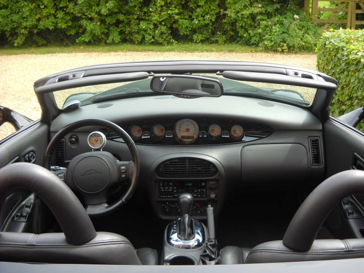 Plymouth prowler - Page 1 - Yank Motors - PistonHeads - The image captures a luxurious scene from inside a sports car. The viewer's eyes are drawn to the black leather interior, including the dashboard which is adorned with multiple dials. The steering wheel, made of a gleaming material, sits on the right side of the car, in accordance with the British driving manual. Through the front window of the car, the outdoors are visible with greenery, suggesting a serene and picturesque surroundings. The car appears to be parked, as indicated by the fact that the engine cover is up. Despite these features, there is no driver visible in the image.