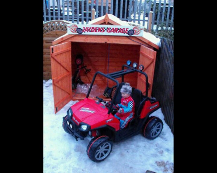 Kids Pistonheads Junior - The image depicts a young child sitting in a pretend toy car designed to resemble a UTV or all-terrain vehicle. The car is small, with a cabin that resembles a genuine UTV's structure. It's parked near a structure resembling a wooden shed or outhouse with a tiered roof, which has red and yellow lettering on it, possibly reading "Jada's Trash." The ground is covered in snow, hinting at a cold season. The child appears to be enjoying the toy vehicle, which adds a playful and whimsical touch to the scene.