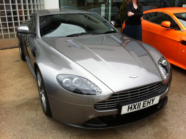 KX60 *** Orange Vantage V12 - North London - Anyone? - Page 1 - Aston Martin - PistonHeads - A silver sports car is parked on a street near a black car. There is a person standing next to the silver car, appearing to be inspecting it. The license plate of the sports car reads "HX11EY". The scene suggests a car show or an auto dealership, where people often go to look at and possibly purchase cars.