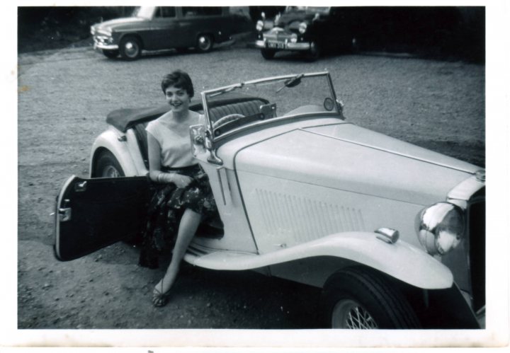 Pistonheads Identify - The image is a black and white photograph of an older car featuring a convertible design. A woman is seated in the open doorway of the car, leaning her elbow on the window and smiling towards the camera. She is dressed in a short-sleeved dress and flat sandals. The car is parked on a gravel or dirt area, with other vehicles partially visible in the background. The overall lighting and quality suggest this photo may be from the mid-20th century.