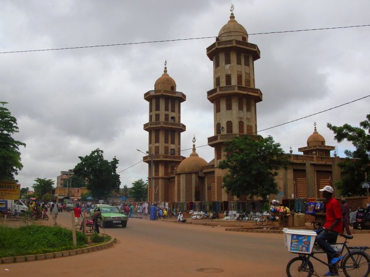 A clock tower in the middle of a city - Pistonheads - The image shows a bustling street scene with a row of tall, brown minarets rising above a large crowd. In the foreground, there's a dark blue bicycle with a white basket on the back, ridden by a man wearing a red shirt. The street is busy with people, vehicles, and shops, and there appears to be a market or bazaar nearby. The minarets have distinctive domed shapes on the top, common in Islamic architecture, and are part of a notable landmark or mosque. The sky is overcast, suggesting an early morning or late afternoon time.