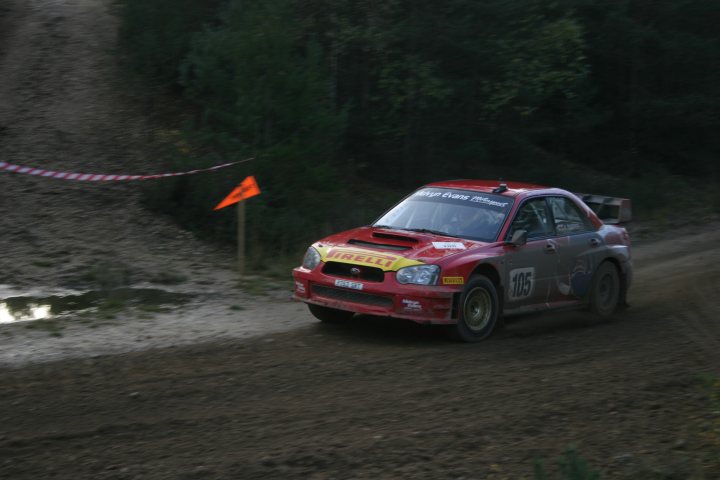 Tempest Rally tomorrow. Anyone on here going? - Page 1 - Thames Valley & Surrey - PistonHeads - In the image, we see a red car with yellow lettering, possibly indicating a racing team or number. The car is caught in the act of navigating a turn on a dirt road, with a tire full of mud spraying out, implying high speed or the road's muddy condition. The car is situated within a dense forest of dark green trees, setting a vibrant contrast against the red of the vehicle. Interestingly, a red flag is visible in the background, which could signify a stop sign or a barrier marking the end of the path for the car.