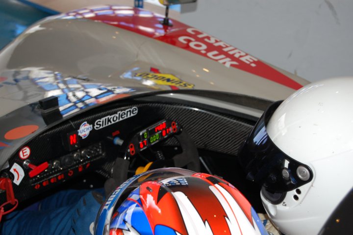 Pistonheads - The image showcases the cockpit of a racing car display, featuring two black car seats mounted on a blue and white baseplate. The right seat has a red, white, and blue helmet nearby, adding a touch of color to the otherwise monochrome setup. At the back, a branded air disruptor is visible, ensuring racers have clean air during high-speed races. The overall arrangement suggests a simulator for racing enthusiasts.