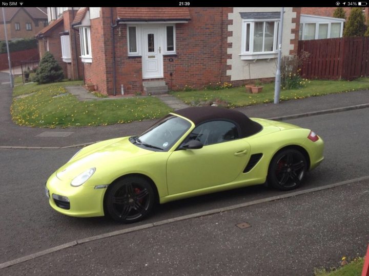 Looking For My Old 'Krypton Green' Boxster... - Page 1 - Boxster/Cayman - PistonHeads - This is a color photograph depicting a shiny, two-door convertible sports car parked on the side of a street. The car appears to be yellow with black tires and trim, and has a black convertible roof that is currently up. The vehicle is positioned close to the edge of the pavement, leaving a small space between it and the curb. The setting is a suburban neighborhood with a sloped driveway leading to a house with a red brick facade and a tiled roof. In the background, the house number is visible, and there's a wooden fence along the property line. The sky is overcast, indicating cloudy weather conditions.