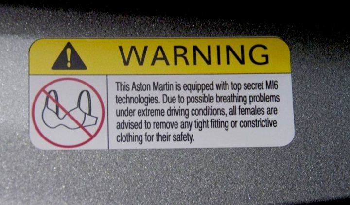 Is it a bad idea to remove this? - Page 1 - Aston Martin - PistonHeads - The image displays a warning label attached to a metal surface. The label is predominantly yellow with red and black text and a black and orange pictogram, commonly recognized representations for restricted or hazardous areas. The text emphasizes that the Aston Martin company has equipped the area with "top secret MI6 technologies" and warns that conditions might lead to extreme driving situations or cause oxygen deprivation. It also advises users to remove personal tight-fitting or constrictive clothing for their safety. The label serves as a cautionary notice, likely intended for a specific group, possibly referred to as the 'MI6' team, and is situated in a place not opened to the public. The background is a beige indoor surface which provides a contrast to the label.