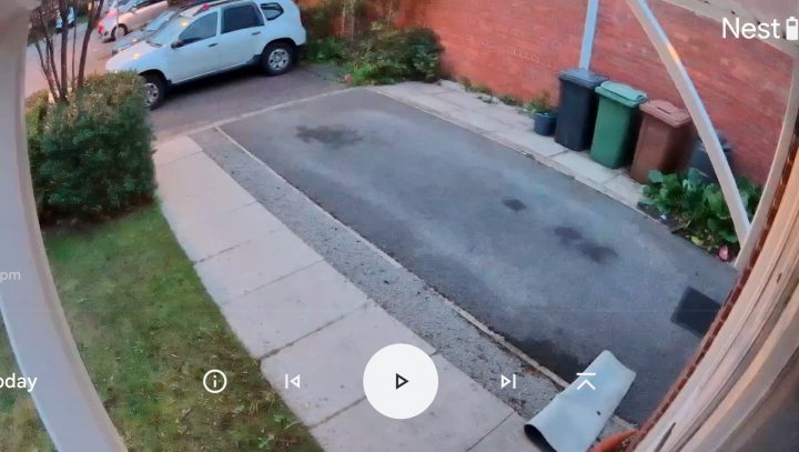 Pistonheads - The image depicts an outdoor scene viewed through a camera lens. It shows a driveway with a small car parked, bordered by brick walls on both sides and a red building visible in the background. A surveillance camera is mounted high on a wall near the entrance of the driveway. The photograph appears to have been taken during the day under overcast skies, as indicated by the grayish tone and the presence of shadows from trees or other structures. There are no people in view, suggesting that the image may have been captured at an unoccupied time. The image quality is somewhat blurry, indicating it might be a still image from a security camera or a screenshot from a video surveillance system.