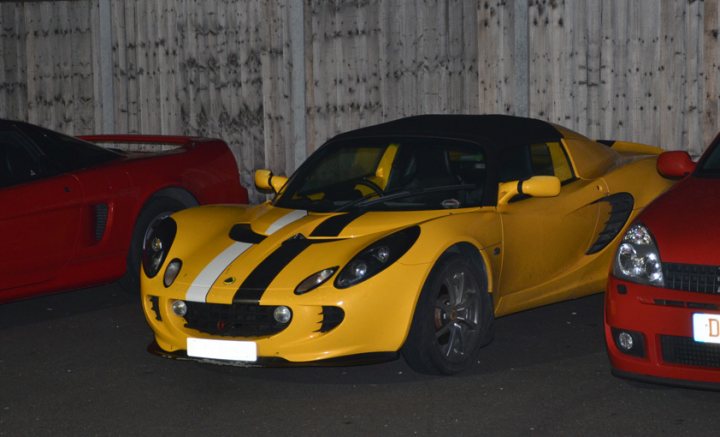 Essex Pistonheads March - The image captures a serene night scene in a parking lot, featuring three cars parked side by side. The first and most striking is a bright yellow sports car, its vibrant color standing out against the darker hues of its companions. Next to the yellow car is a sleek red sports car, its glossy exterior reflecting the night lights. The third car, parked in line with the other two, is a more traditionally colored black sedan. The cars are parked in front of a sturdy-looking stone wall, which serves as a backdrop to the scene. There is no sign of human activity, giving the image a calm and still ambiance.