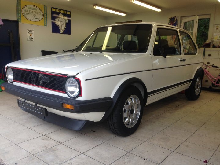 VW Golf GTi MK1 Restoration  - Page 9 - Audi, VW, Seat & Skoda - PistonHeads - The image shows a classic white Volkswagen Beetle car in an indoor setting, likely a garage or a showroom. The car's distinctive rounded body is visible, with the black grille framing the front. The wheels feature a silver hubcap, and the vehicle appears to be in good condition, without visible damage or scratches. The car's license plate is also visible, though the specific registration number is not clear. In the background, there are banners and a sign that reads "Michelin" and others that are too distant to decipher.