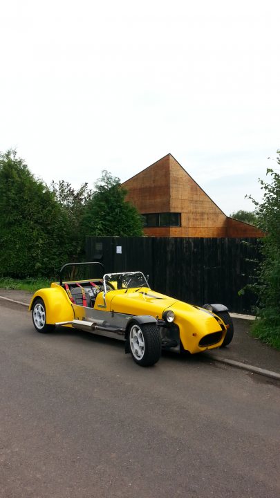 Who's got what in the South West - Page 4 - South West - PistonHeads - The image shows a single yellow Chopper parked on the side of a road. The photo appears to be taken during the day under an overcast sky. The chopper is positioned parallel to the curb of the street, and there is a sturdy wooden fence visible behind it. In the background, there is a building and lush green trees, blending a touch of urban and natural elements. The chopper's design is sleek and modern, featuring a stretched-out frame and a minimalist aesthetic.