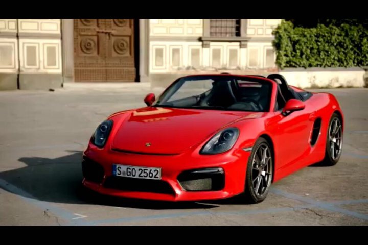 GT4 colours  - Page 52 - Boxster/Cayman - PistonHeads - The image is a photograph of a high-end sports car, which appears to be a Porsche, given its distinct design features and the model badge. The vehicle is a convertible with a folding roof that is currently stowed beneath the rear windshield. It's colored in a vibrant red hue, showing a glossy finish that reflects some of the surrounding environment. The car is parked on a concrete surface, possibly a parking lot, in front of a building with classical architecture. There is no text present in the image. The sky is overcast, and the atmosphere suggests partially cloudy conditions.